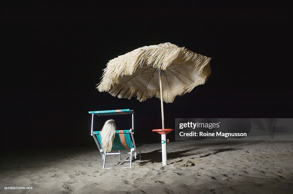 Woman sitting on deck chair on beach at night, rear view