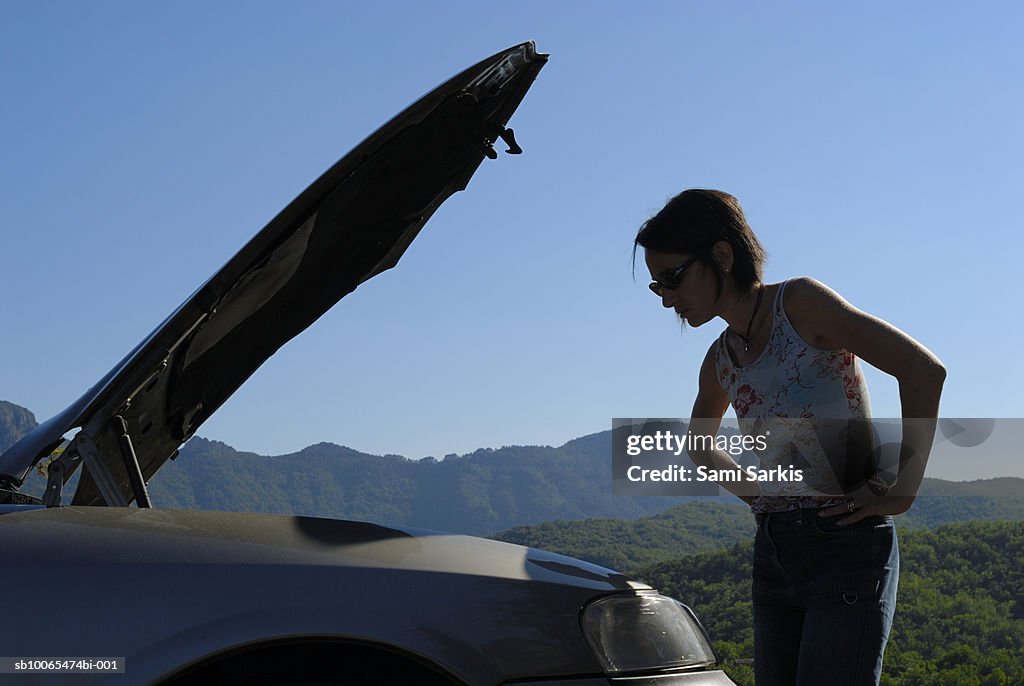 Woman with broken car in rural setting, looking at engine, side view