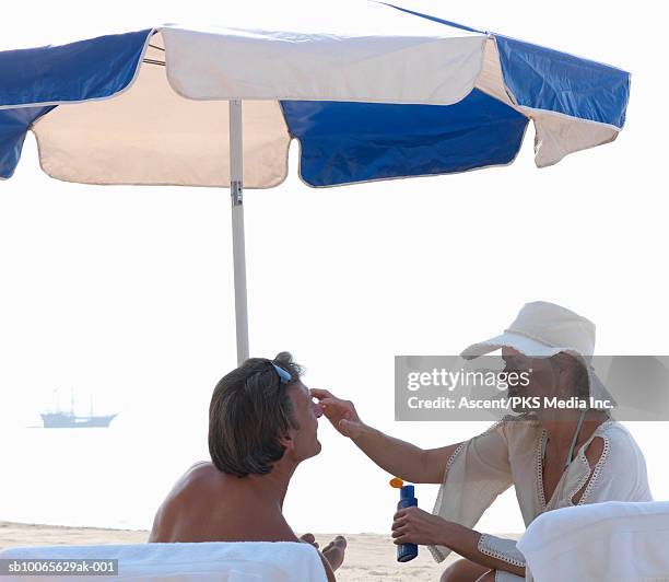 mature couple on beach, woman applying sun cream to man's nose, smiling - cream colored hat stock pictures, royalty-free photos & images