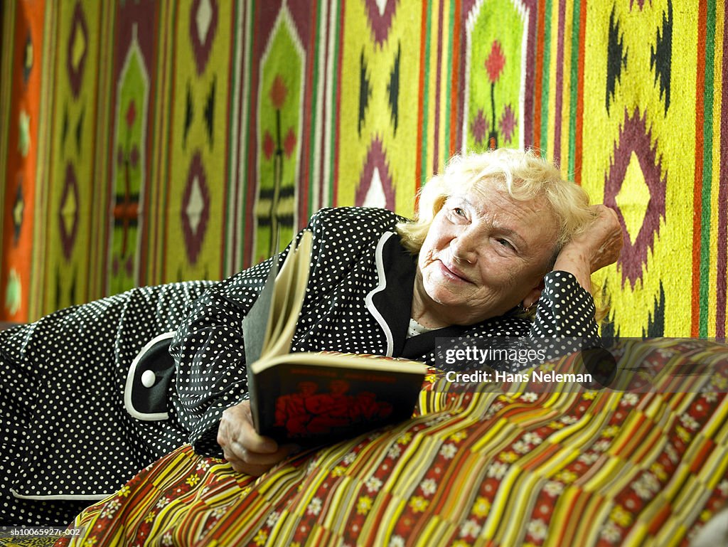 Senior woman reclining on bed holding book, smiling