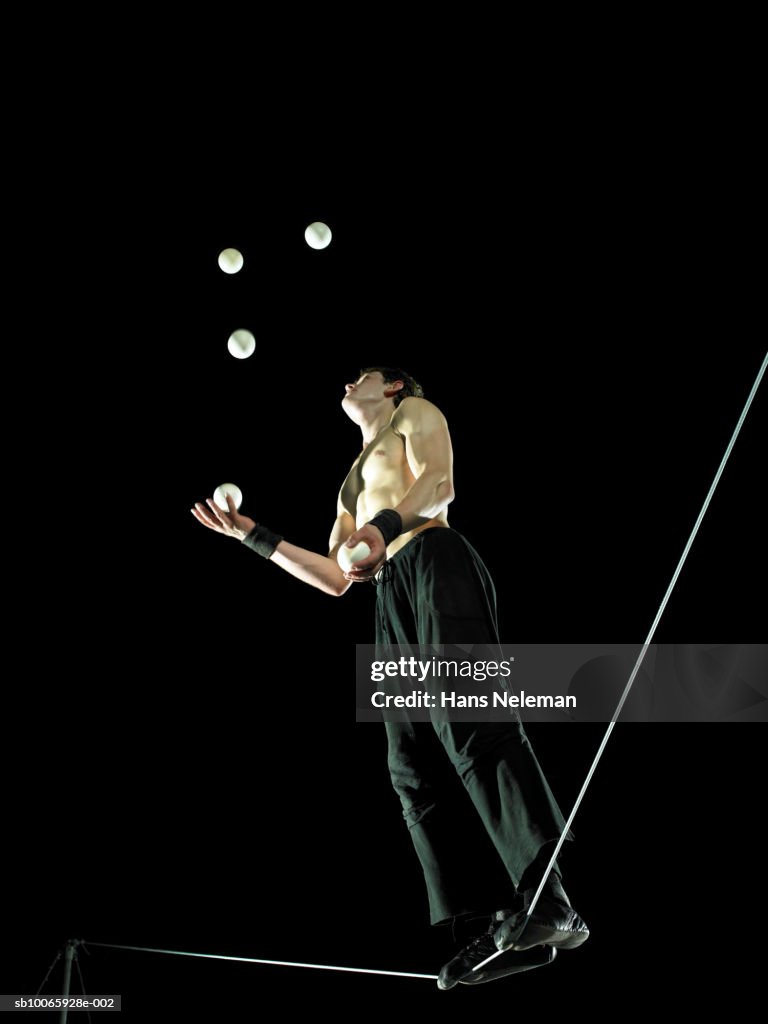 Male gymnast standing on tightrope, juggling balls, low angle view
