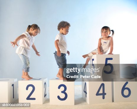 Baby boy (21-24 Months) and two girls (2-5) climbing on lettered white boxes Baby boy (21-24 Months) and two girls (2-5) climbing on lettered white boxes