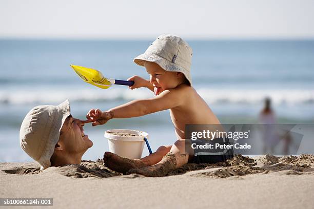 father buried in sand and young boy (2-3) with bucket and spade on beach - バケツとスコップ ストックフォトと画像