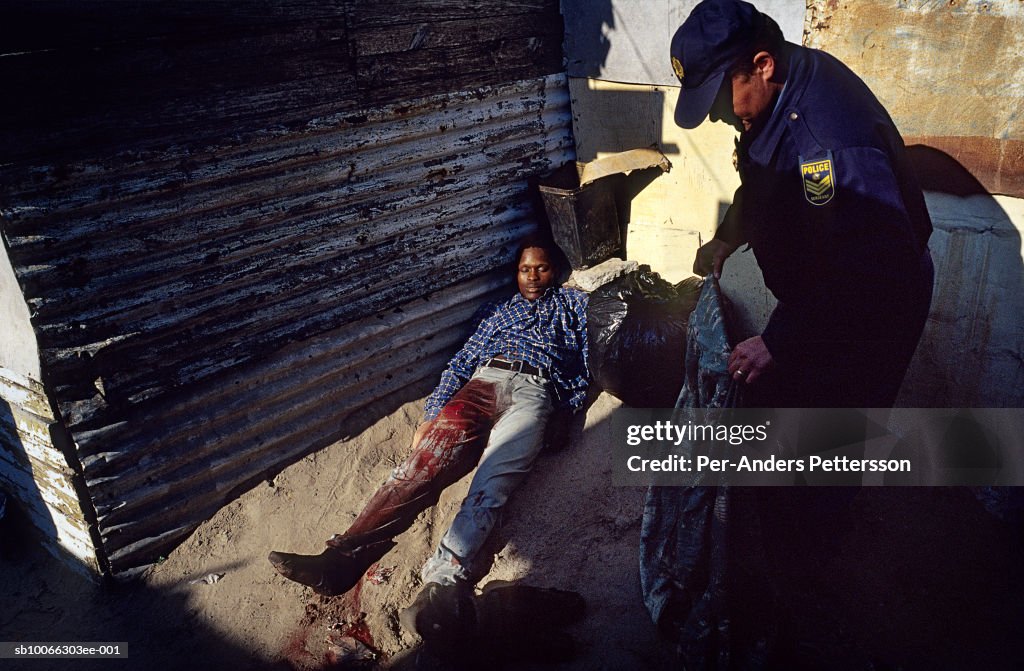 South Africa, Cape Town, Khayelitsha, police officer places blanket over dead man outside shack