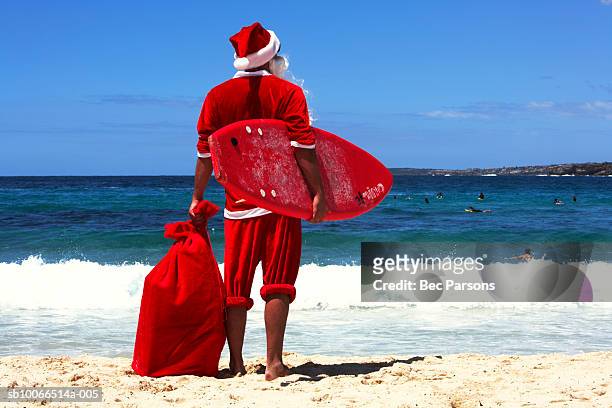 man dressed as santa claus holding sack and surf board standing on beach, rear view - out of context imagens e fotografias de stock