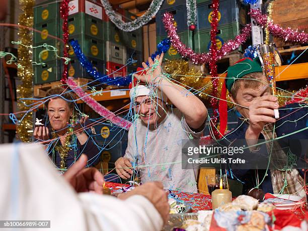 three people covered in party streamers laughing at charismas table in warehouse - adult laughing christmas stock pictures, royalty-free photos & images