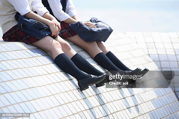 two female high school students (17-18) sitting on top of tiled wall, low section - japanese short skirts stockfoto's en -beelden