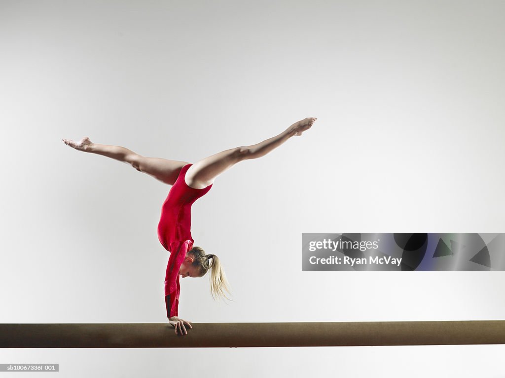 Teenage gymnast (15-16) doing handstand on balance beam, studio shot