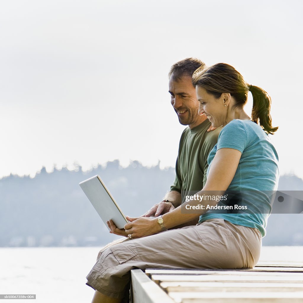 Mature couple using laptop on edge of pier, smiling, side view