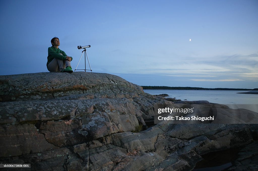 Woman with telescope sitting on seashore at dusk