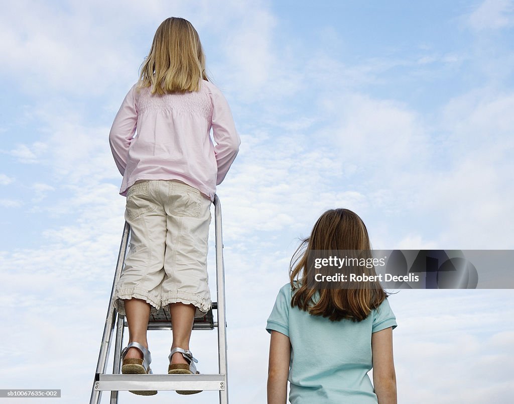 Girl (8-9 years) standing next to sister (6-7 years) standing on ladder, rear view