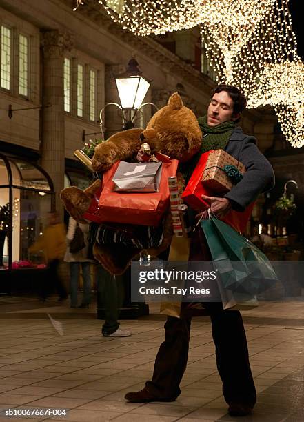 man holding shopping bags and giant teddy bear, standing on street at night - men stock photos et images de collection