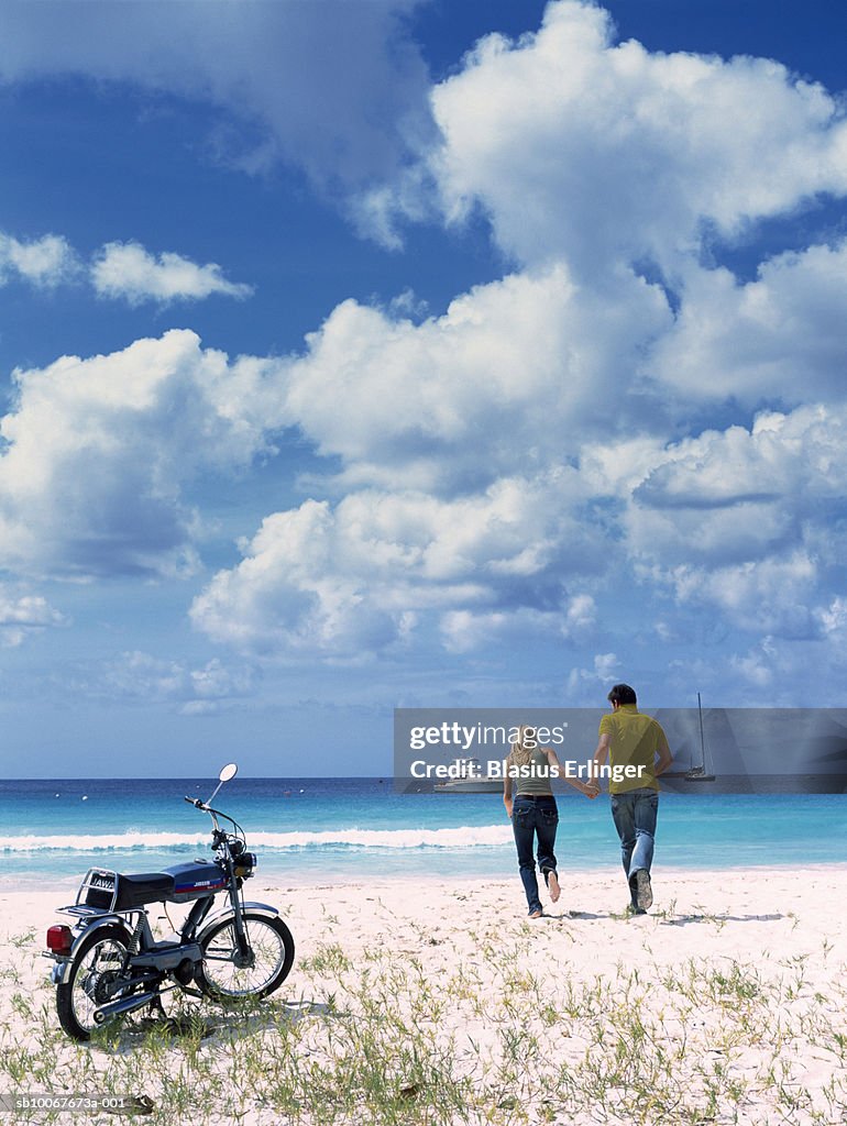 Couple holding hands running towards ocean, motorbike in foreground, rear view