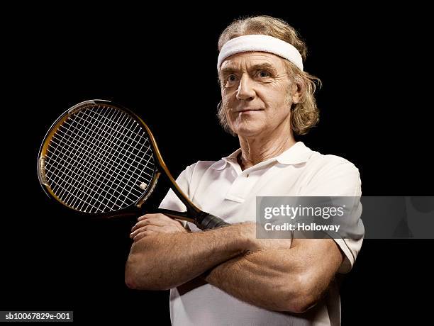 mature man with arms crossed holding tennis racket on black background - cinta deportiva del pelo fotografías e imágenes de stock