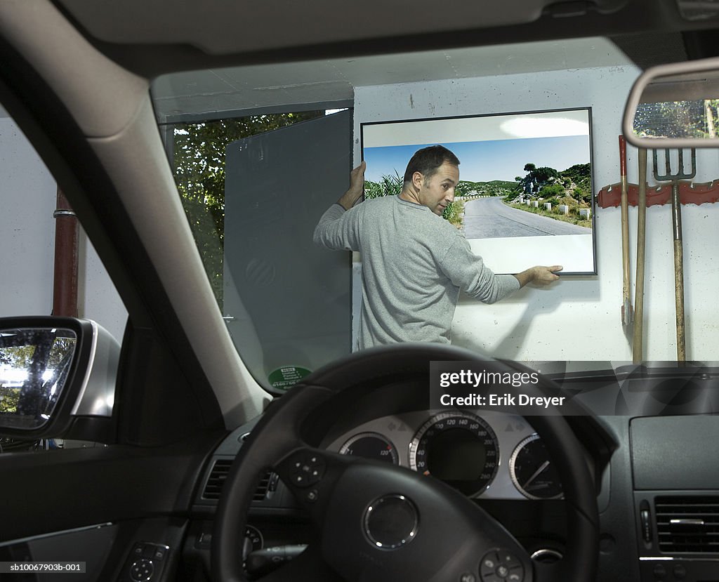 Man hanging picture on wall in garage, view from car interior