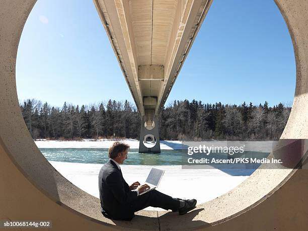 businessman using laptop on circular pillar of bridge, frozen river in background, side view - calgary bridge stock pictures, royalty-free photos & images