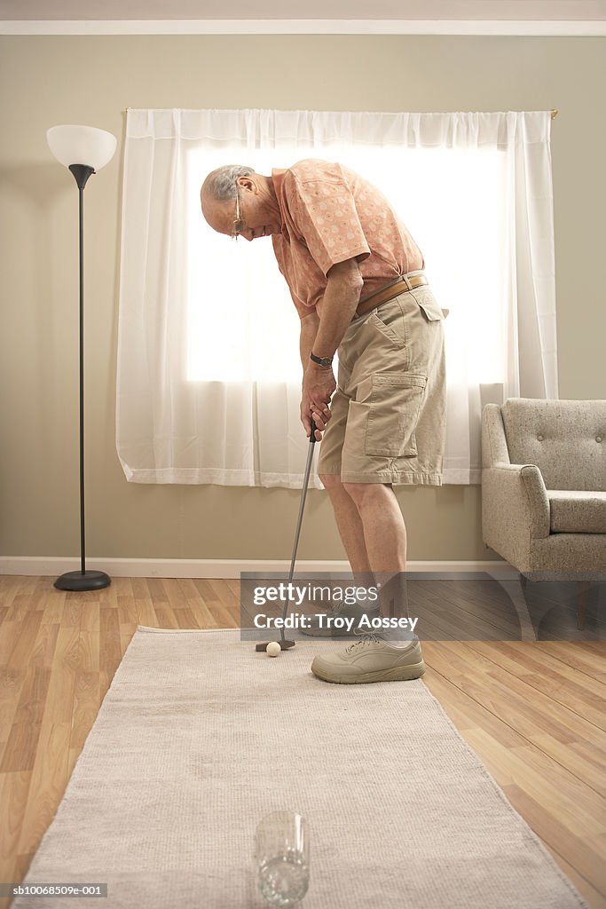 Senior man in living room playing golf, putting into glass, side view