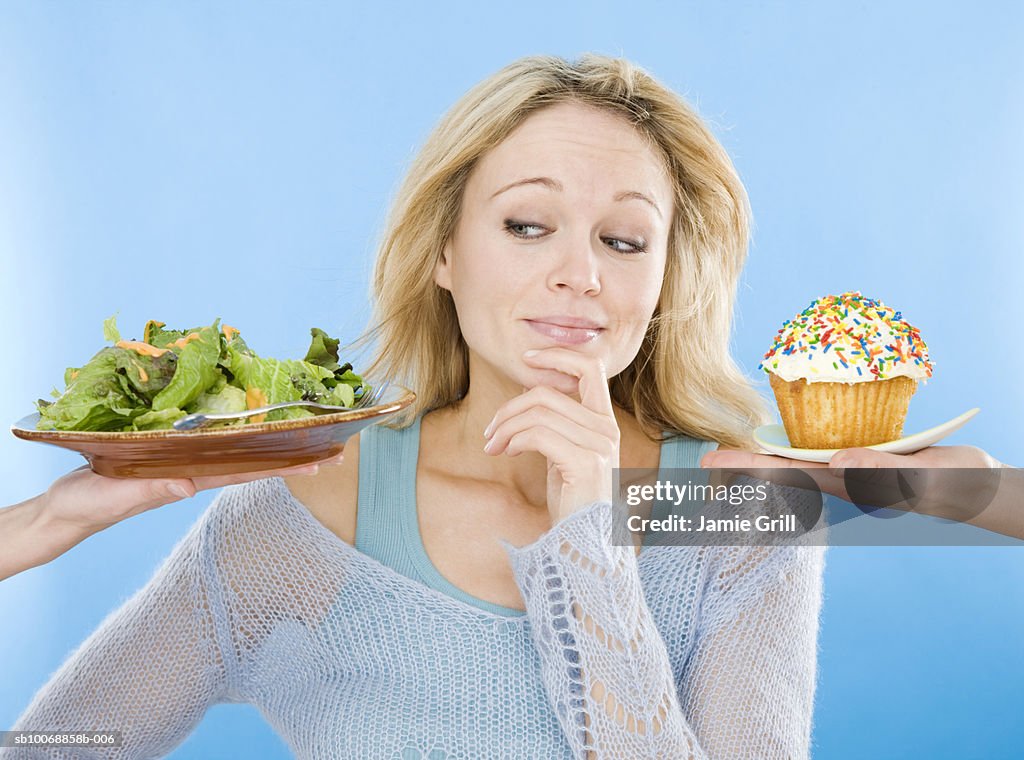 Young woman looking at salad and cupcake, hand on chin, close-up