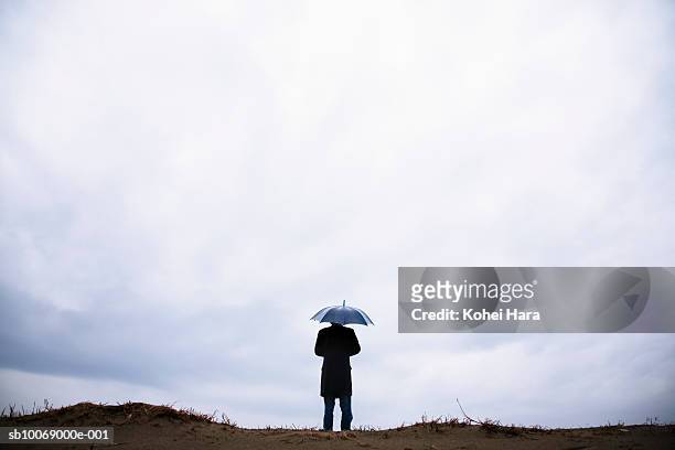 young man standing on small hill holding umbrella, rear view - overcoat stock pictures, royalty-free photos & images
