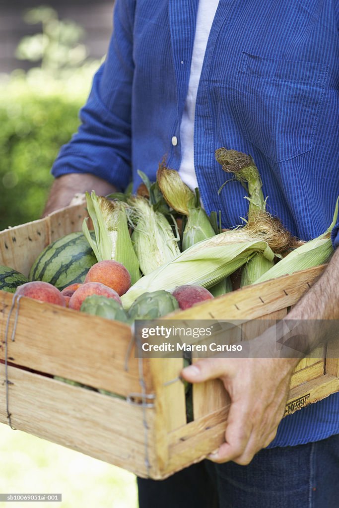 Man carrying wooden box filled with corn in husks, peaches and watermelon, close-up, mid section