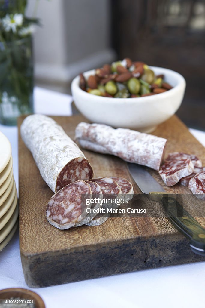 Sliced salami on cutting board with knife, bowl of olives in background, close-up