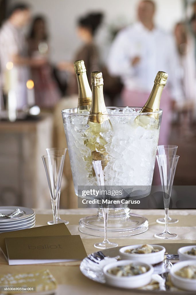 Champagne bottles in ice bucket with glasses, people in background.