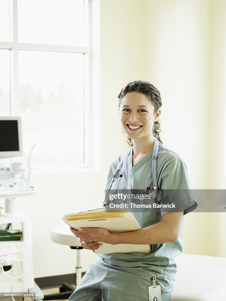 Female nurse in exam room holding file, smiling, portrait