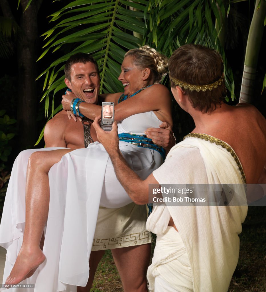 Man carrying woman, friend photographing with mobile phone, laughing
