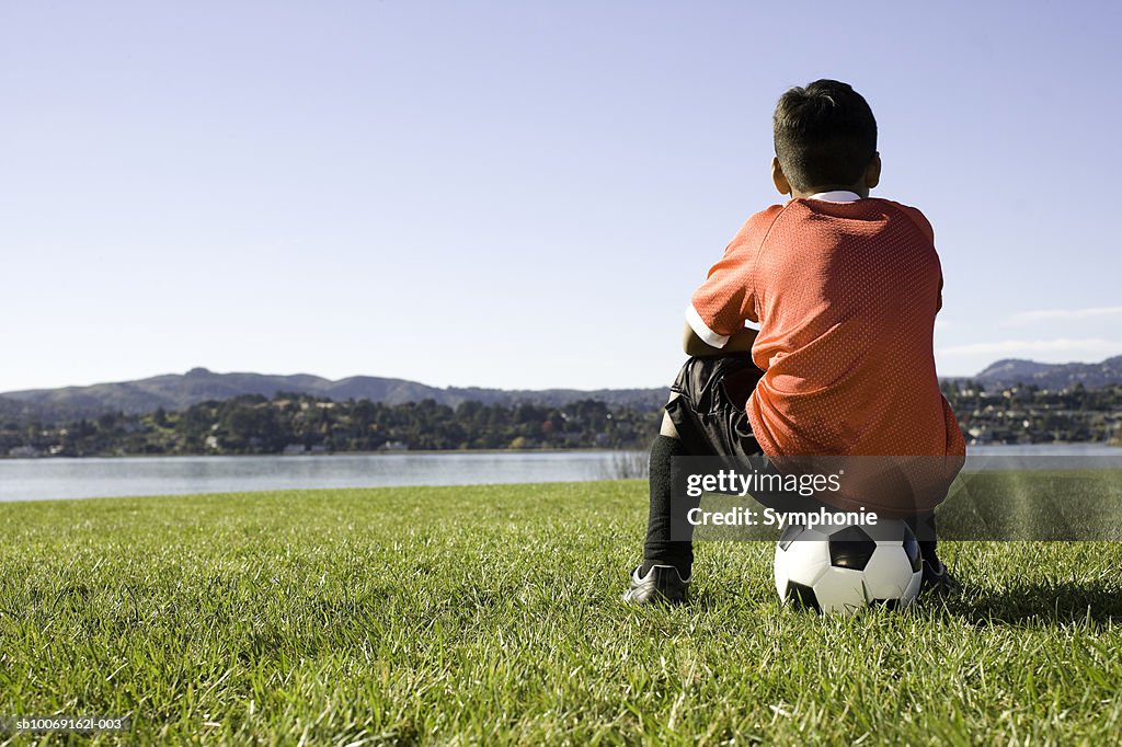 Boy in soccer uniform sitting on ball, rear view