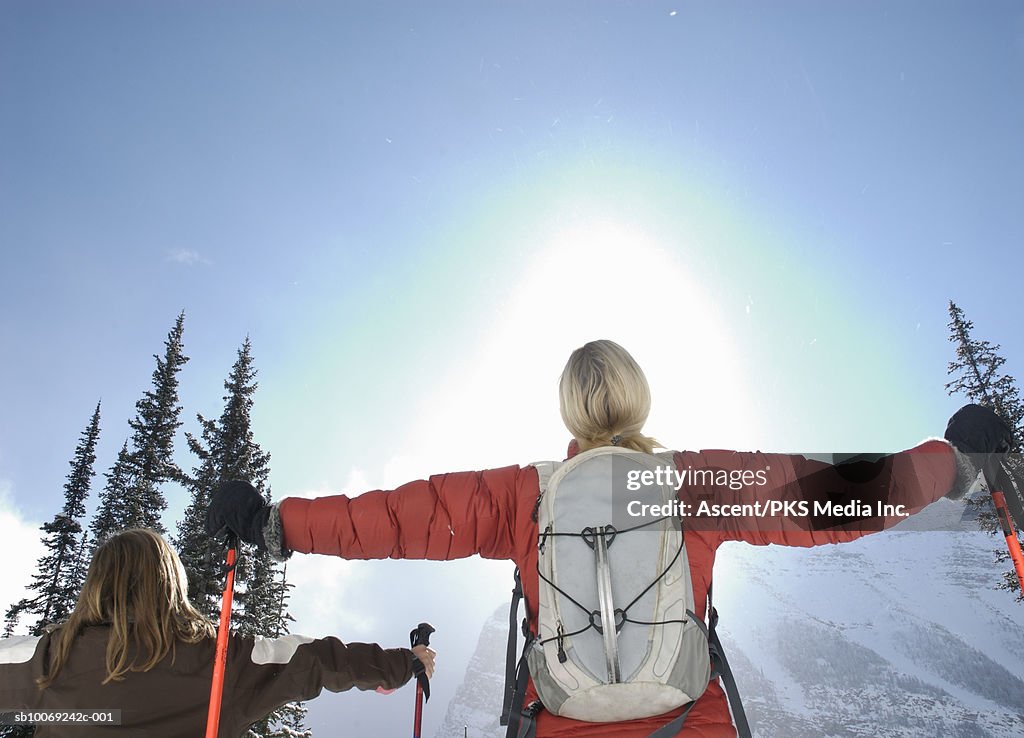 Girl (12-13) and mother holding ski poles looking at sun, arms out, rear view