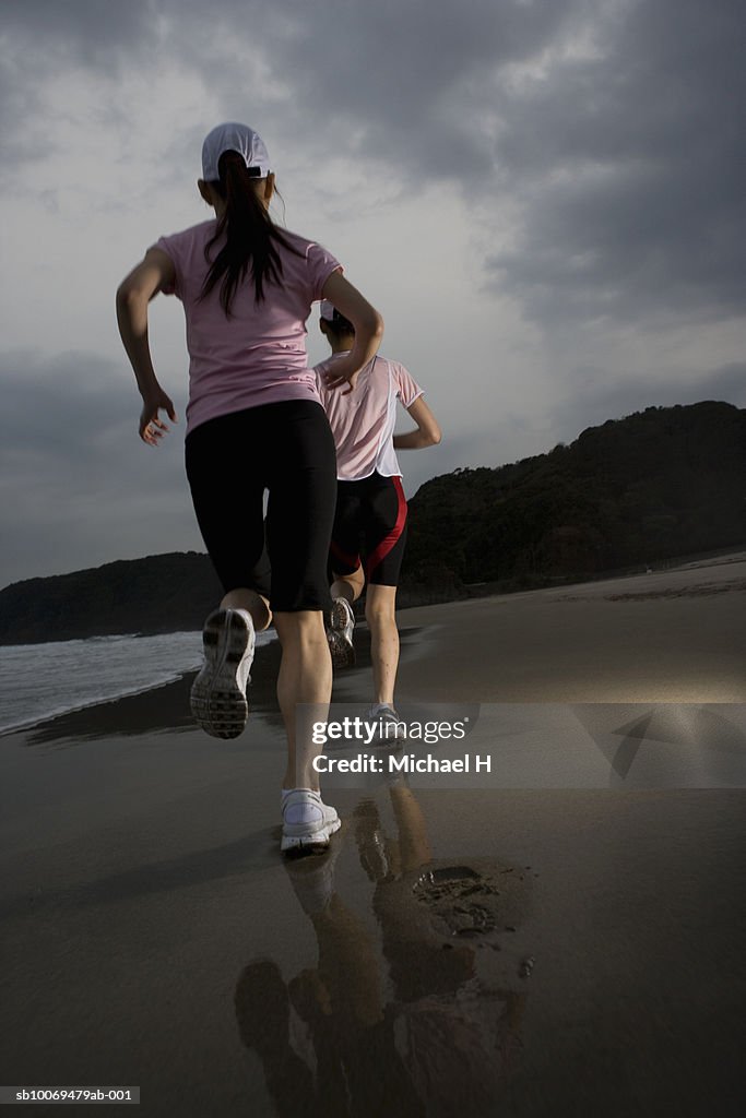 Two women jogging on wet sand at dusk, rear view