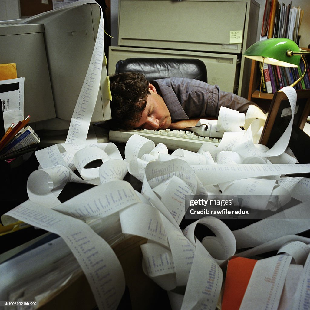 Man sleeping on desk in office with adding machine paper spread all around
