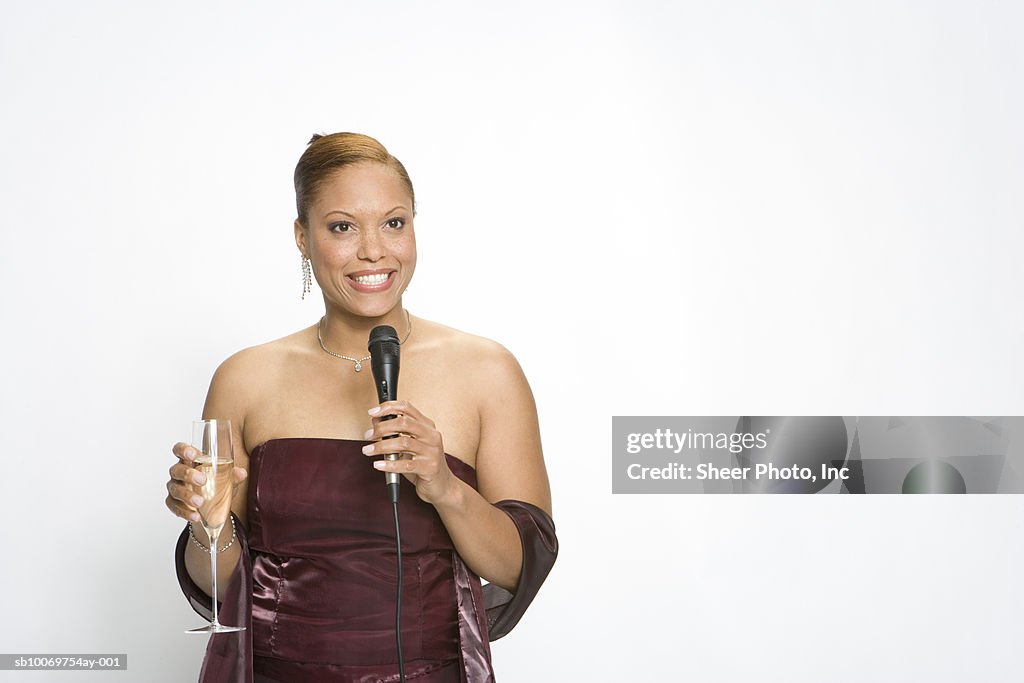 Woman in gown drinking champagne and holding microphone on white background
