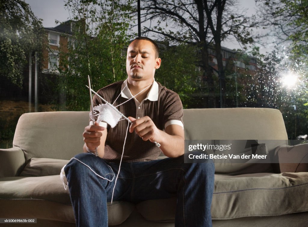 Young man knitting on sofa in living room