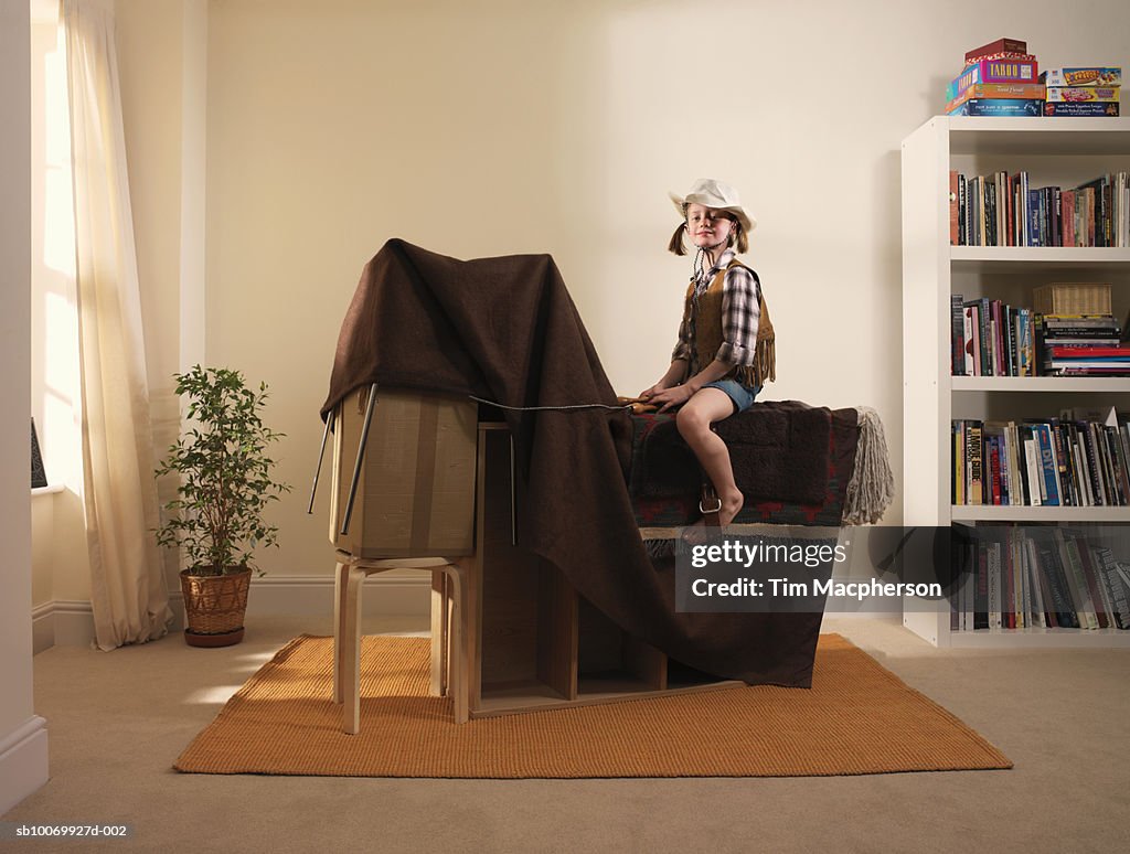 Girl (8-9) pretending to ride horse made from furniture and blanket