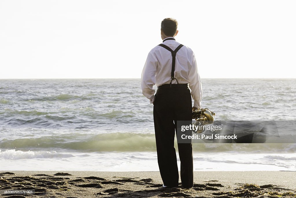 Man with saxophone on beach, facing ocean, rear view