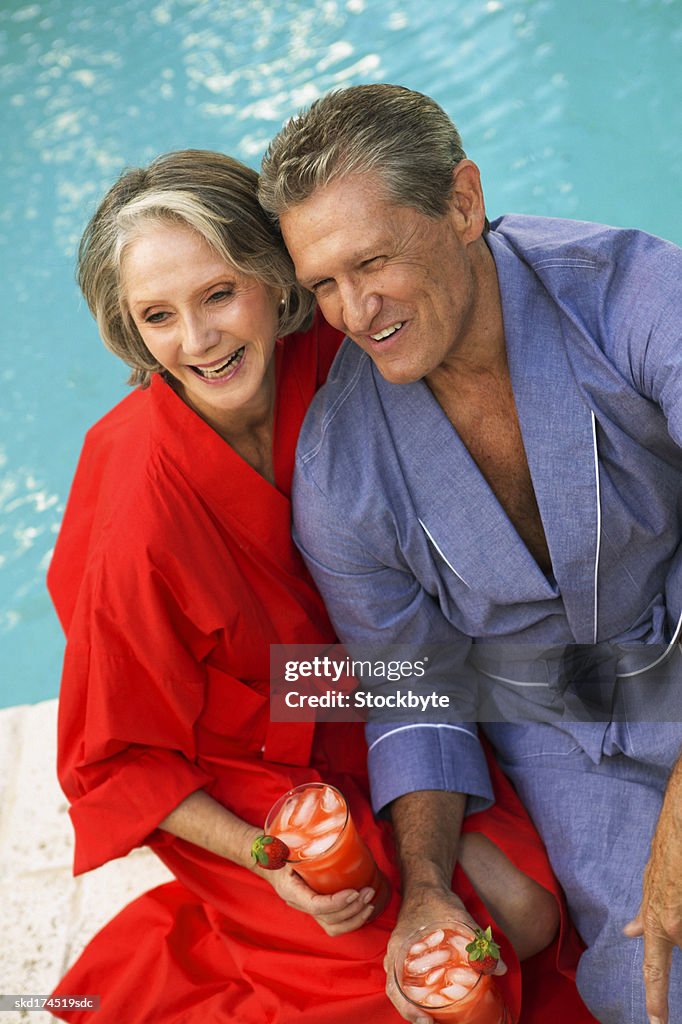 Couple holding cocktails sitting by swimming pool, elevated view