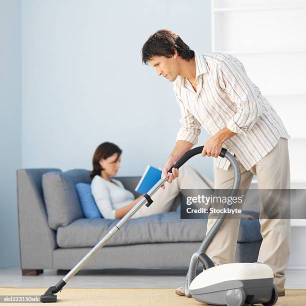 man vacuuming while woman relaxes on a sofa - renversement des rôles photos et images de collection