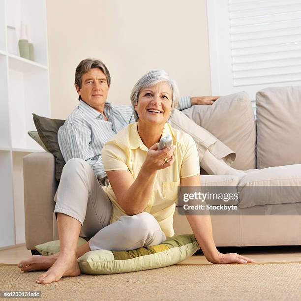 mature couple sitting in living room with woman holding remote control at television - control photos et images de collection