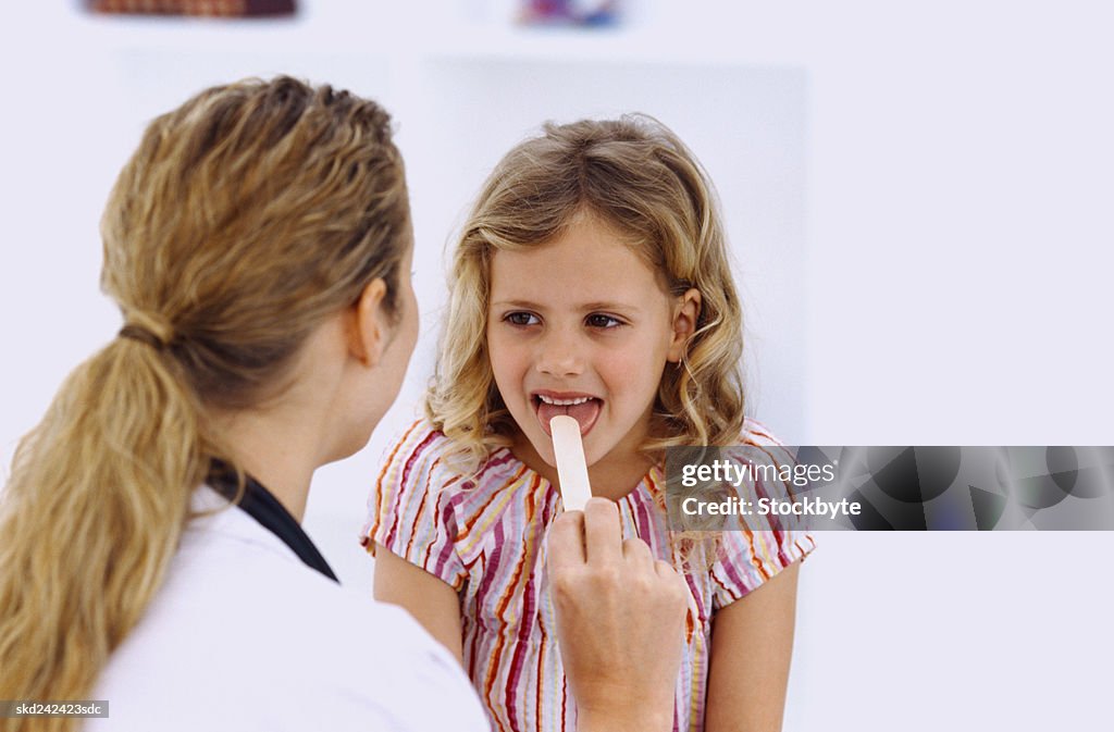 Rear view of a female doctor using a tongue depressor on a young girl (6-8)