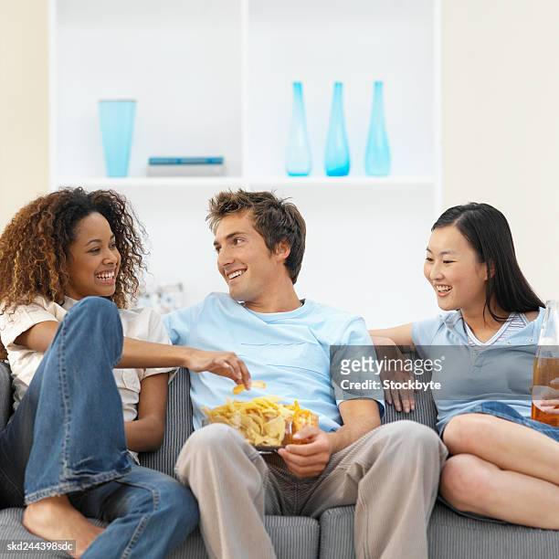 close-up of three young people sitting on sofa and eating crisps and drinking soda - banknoten stockfoto's en -beelden