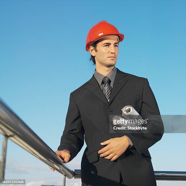 front view portrait of businessman wearing hard hat and holding blueprint under his arm - hat stock-fotos und bilder
