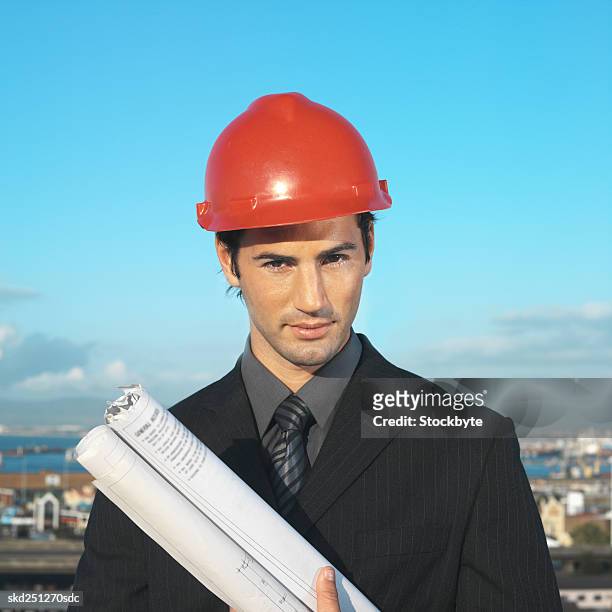 front view portrait of businessman wearing hard hat and holding blueprint - hat stock-fotos und bilder