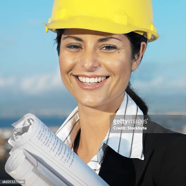 front view portrait of businesswoman wearing hard hat and holding blueprint - hat stock-fotos und bilder