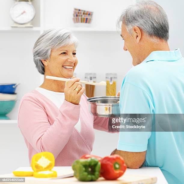 close-up of mature woman feeding mature man spoon of sauce from saucepan with cutting board and bell peppers in front of them - bell fotografías e imágenes de stock
