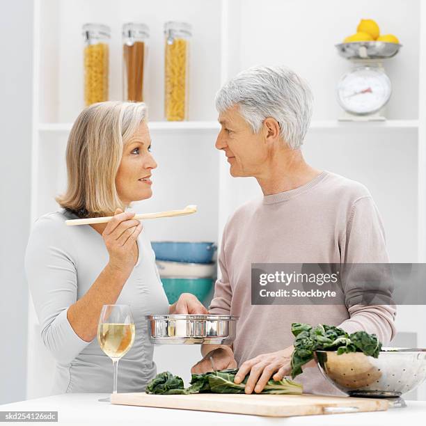 close-up of mature woman feeding mature man spoon of sauce from saucepan with young man cutting cabbage - savory sauce fotografías e imágenes de stock