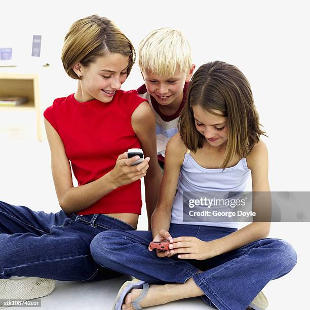 two girls (10-15) using mobile phones with a boy (6-7) looking over their shoulders - 1015 stockfoto's en -beelden