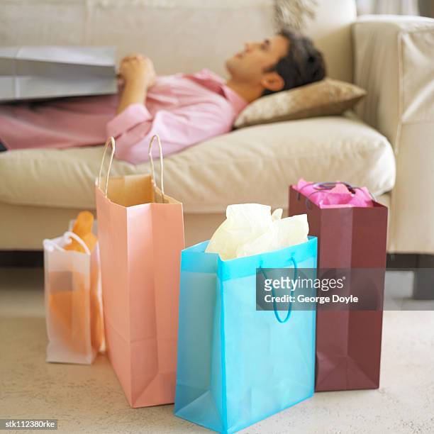 young man lying on a couch next to multicolored shopping bags - next stockfoto's en -beelden