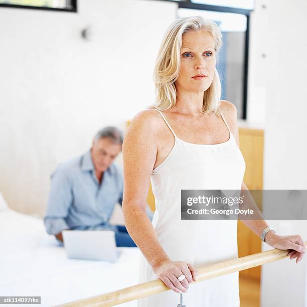 an elderly woman standing against a wooden hand rail and an elderly man sitting with a laptop on a bed behind her - couple 50 55 laptop stock-fotos und bilder