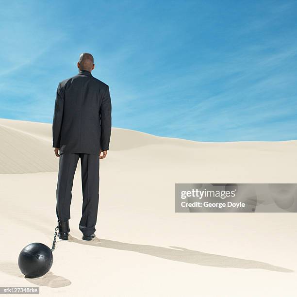 businessman wearing ball and chain in desert rear view, low angle view - restraining device stock pictures, royalty-free photos & images